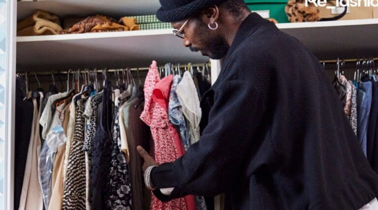 Man in denim jacket and bobble hat looks through a wardrobe of clothing.