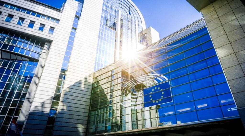 Outside the European Commission, sun shining on tinted glass windows reflecting European flag.