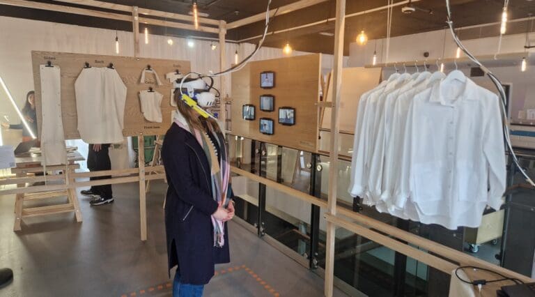 Helena stands in front of rail of redesigned shirts while wearing a VR headset and exploring the future of fashion