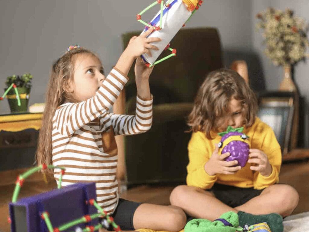 two children playing with toys, one girl is holding a toy rocket above her head while another child sits holding a purple coloured fruit toy.