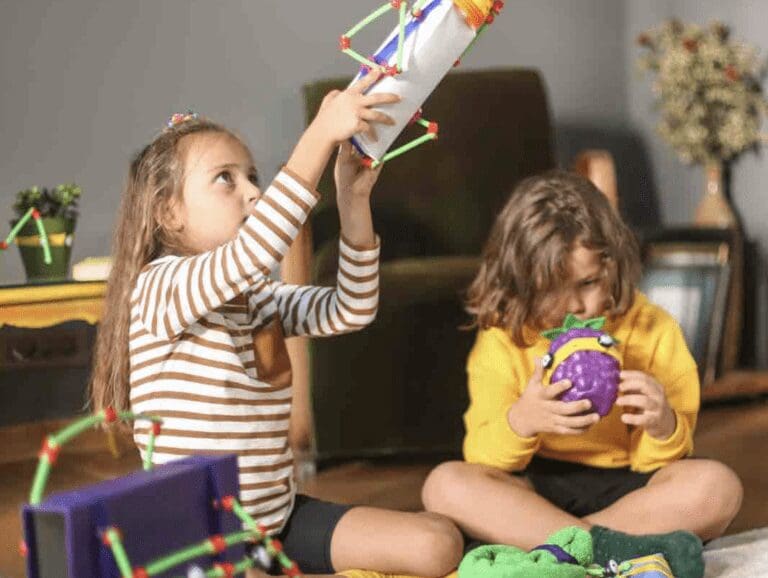 two children playing with toys, one girl is holding a toy rocket above her head while another child sits holding a purple coloured fruit toy.