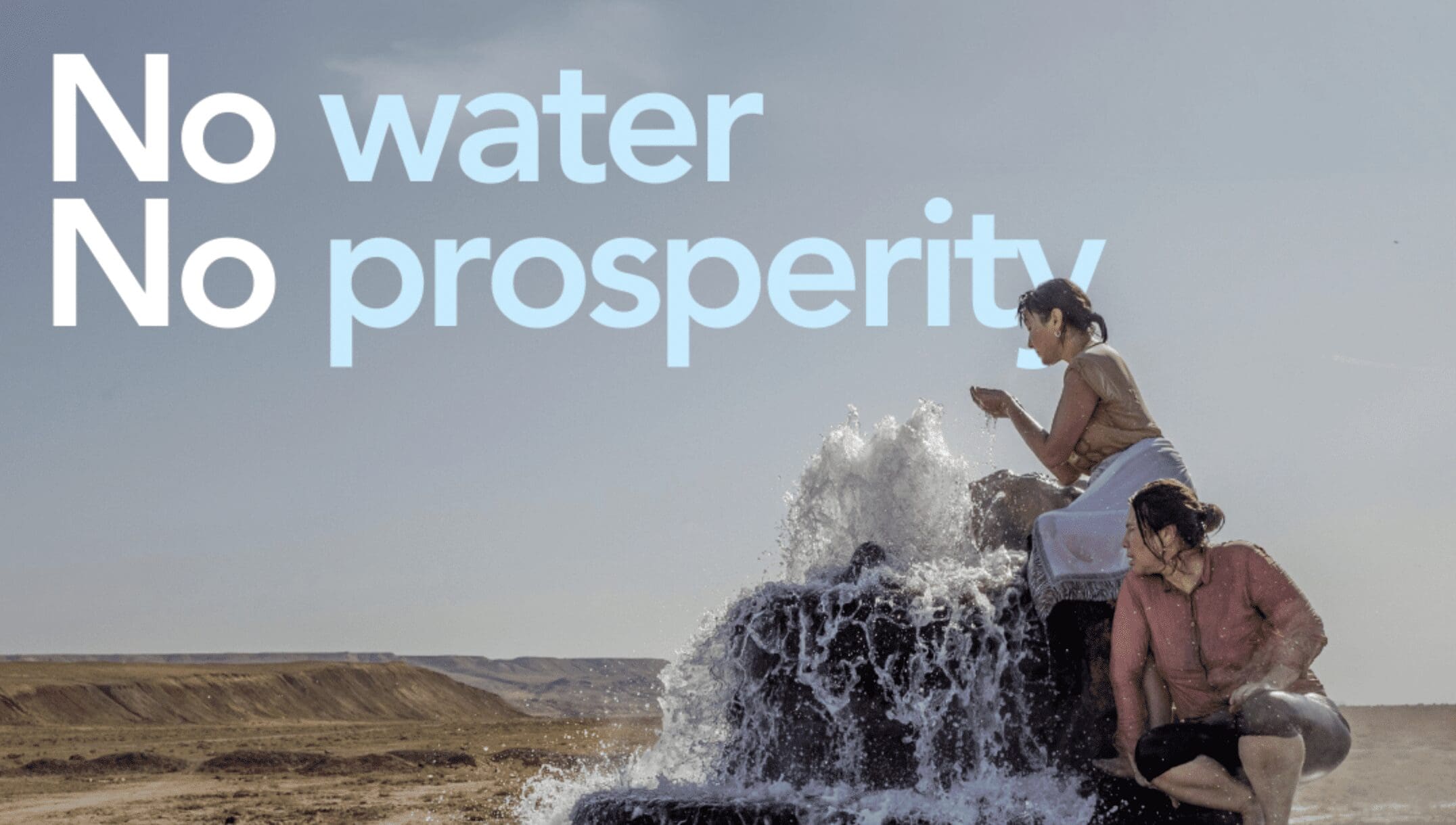 Two women sit on a water fountain in the middle of the desert