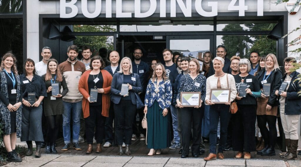 A group of people smile together outside a building as they hold awards certificates.
