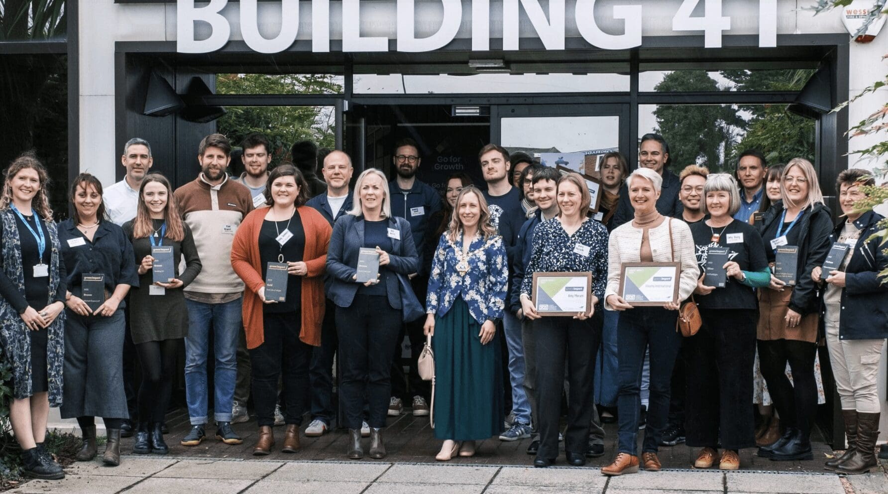 A group of people smile together outside a building as they hold awards certificates.
