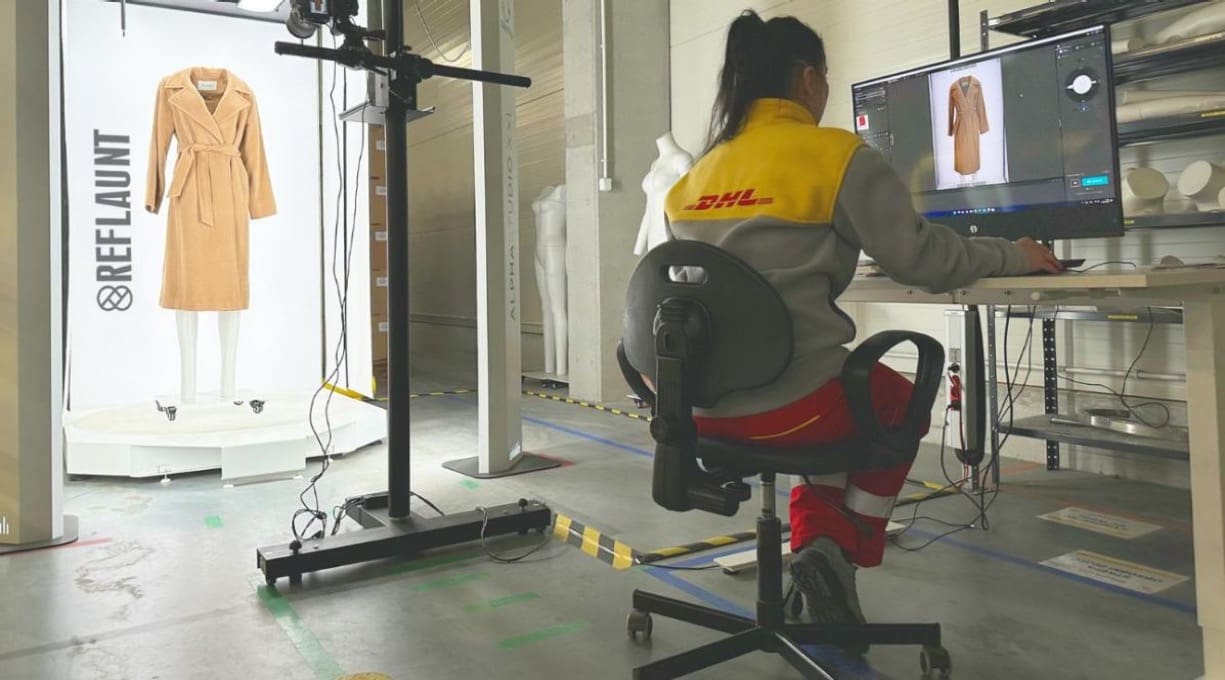 A DHL worker sits at a computer assessing a pre-loved coat on the screen while the coat adorns a mannequin next to her.