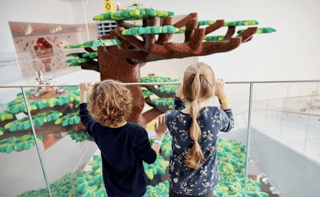 two children peer over a handrail at a large LEGO brick tree.