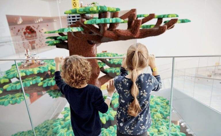 two children peer over a handrail at a large LEGO brick tree.