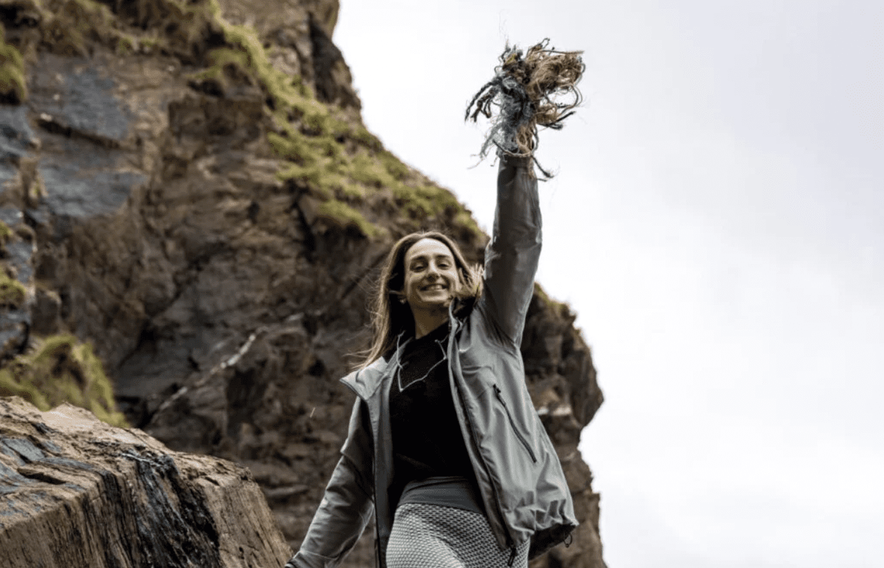 A young woman stands on a shoreline smiling while holding discarded fishing gear up in the air