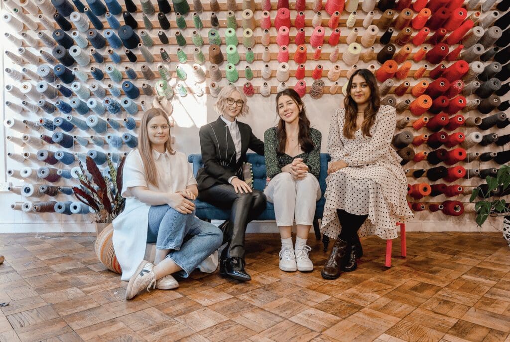 a team of four women sit in front of a wall of cotton and threads in different bright colours