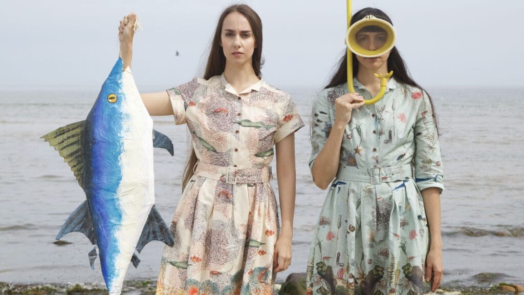Two women stand in front of the sea shore in printed dresses, one wearing a snorkel and mask, the other holding up a fish