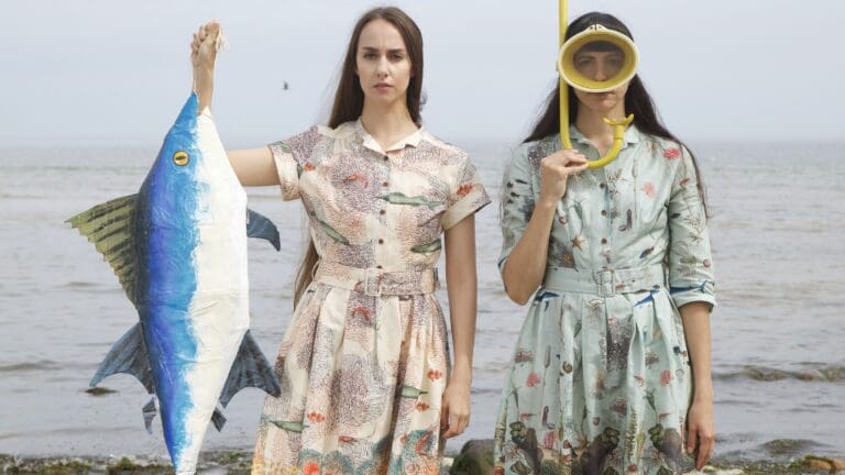 Two women stand in front of the sea shore in printed dresses, one wearing a snorkel and mask, the other holding up a fish