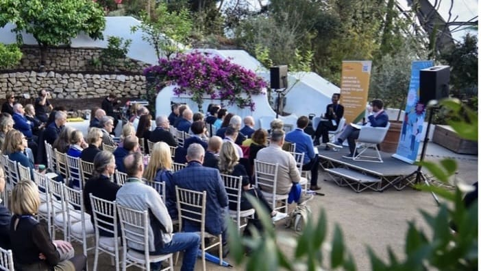 People sat on chairs in front of a stage in a greenhouse